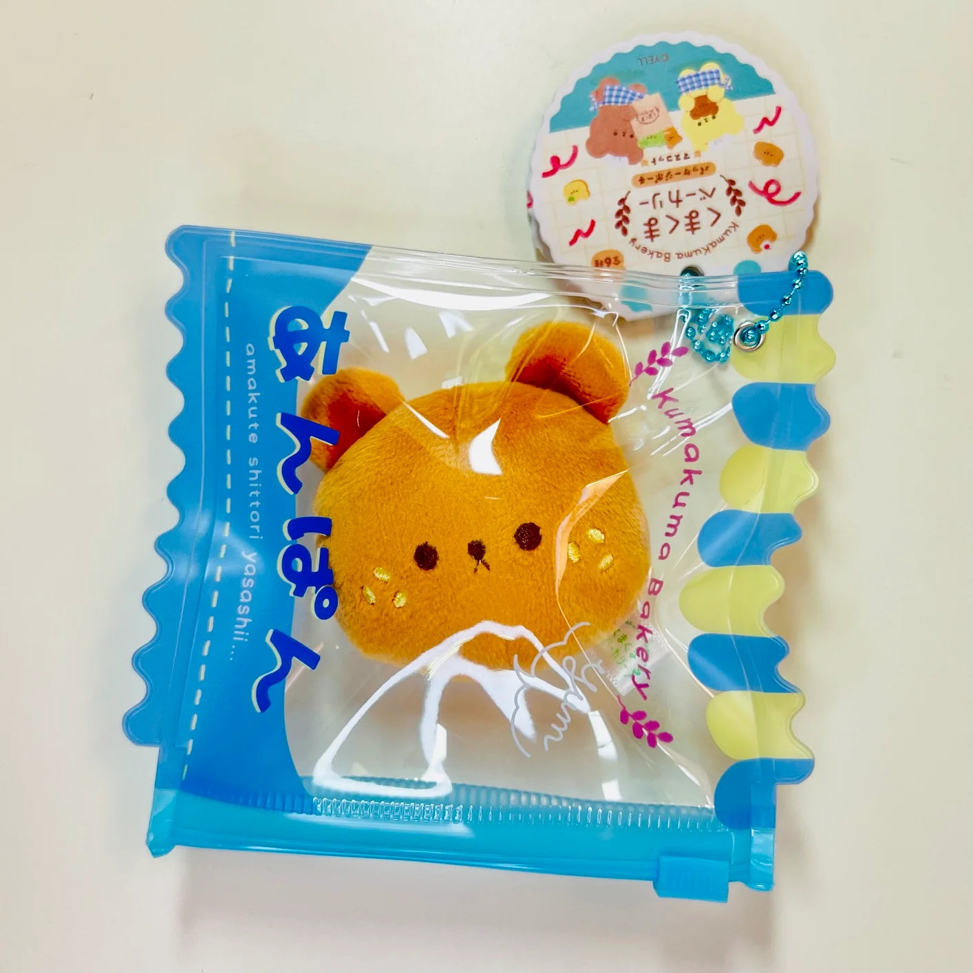 Say hello to the cutest little snack to carry your essentials! This unique Kuma Bakery Bear in a Bag Keychain, Sesame Bun features a soft, squishy bread bear plushie tucked inside a charming clear and blue mini wallet. The softness is like a mochi dessert and the design is unmatched.